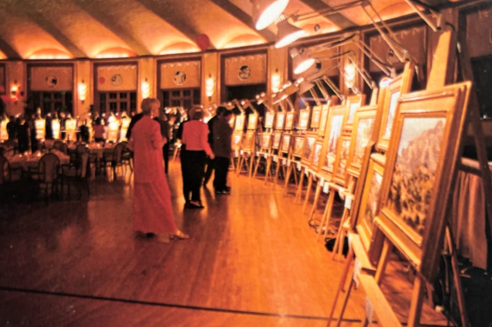 Guests in evening wear viewing a long row of framed paintings on lit easels in the Casino ballroom